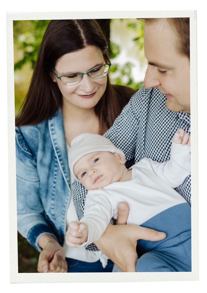 Lifestylowa, naturalna sesja rodzinna w plenerze w parku, wśród drzew. Uśmiechnięta kobieta z brązowymi włosami w okularach w dżinsowej kurtce z mężczyzną w kraciastej koszuli trzymającym na rękach trzymiesięcznego syna w białej koszulce, białej czapeczce i niebieskich spodenkach.